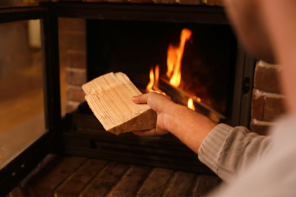 A person holds a piece of light-colored firewood in front of a burning fireplace with a brick surround.