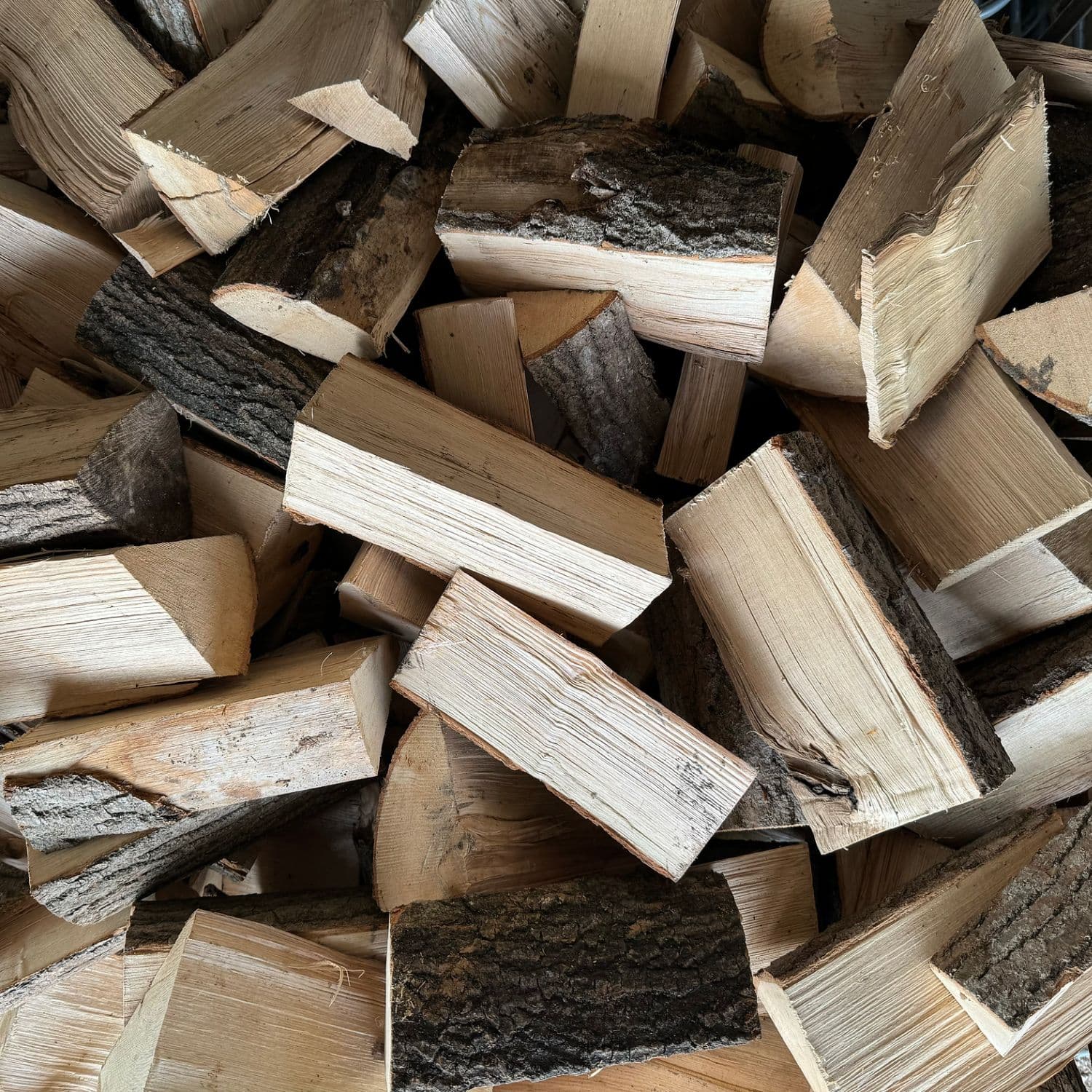 A pile of hardwood firewood logs and split with visible natural bark, light tan wood grain, and darker brown sections.